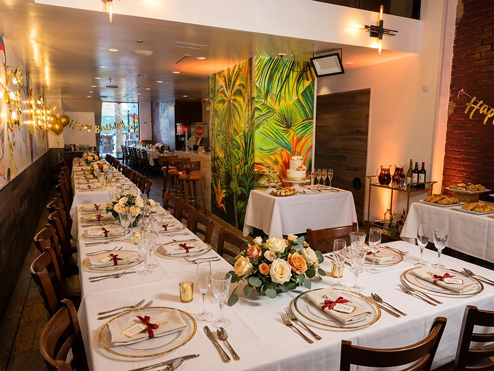 A birthday celebration set with long linen tables, gold chargers, florals and a cake table by the palm mural