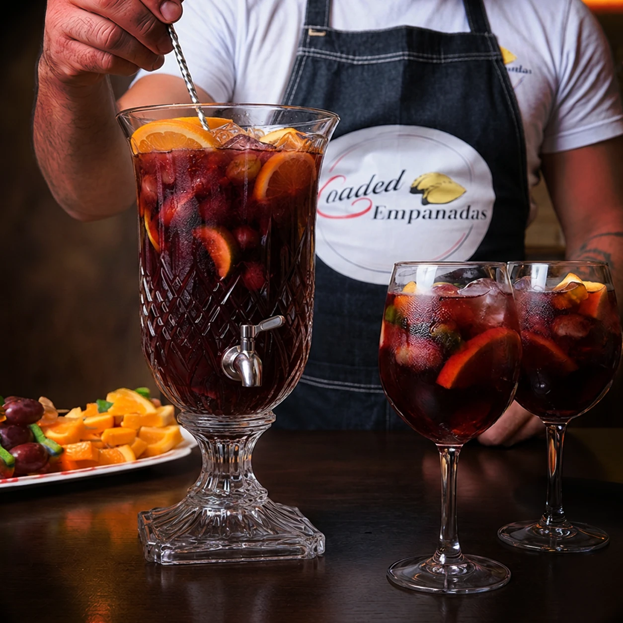 A bartender in a Loaded Empanadas apron stirring a crystal pitcher of red sangria loaded with orange, plum and grapes, two filled glasses alongside