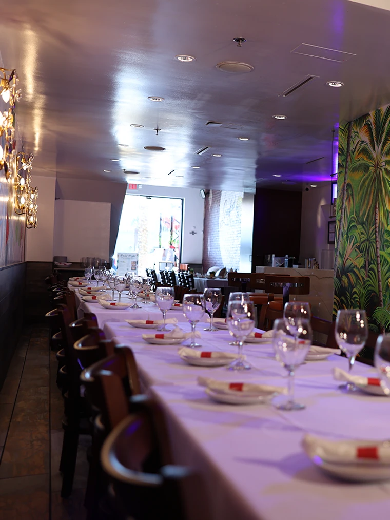 A long banquet table set with white linen, wine glasses and folded napkins beside the palm mural