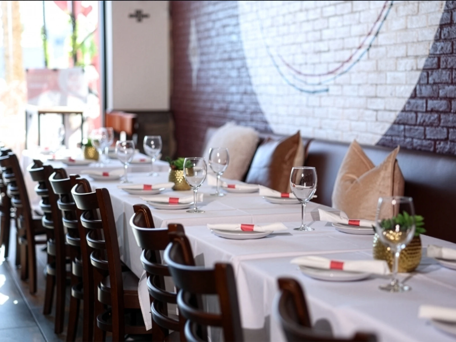A long shared table set with white linens, red-banded napkins and gold centerpieces along the mural wall banquette