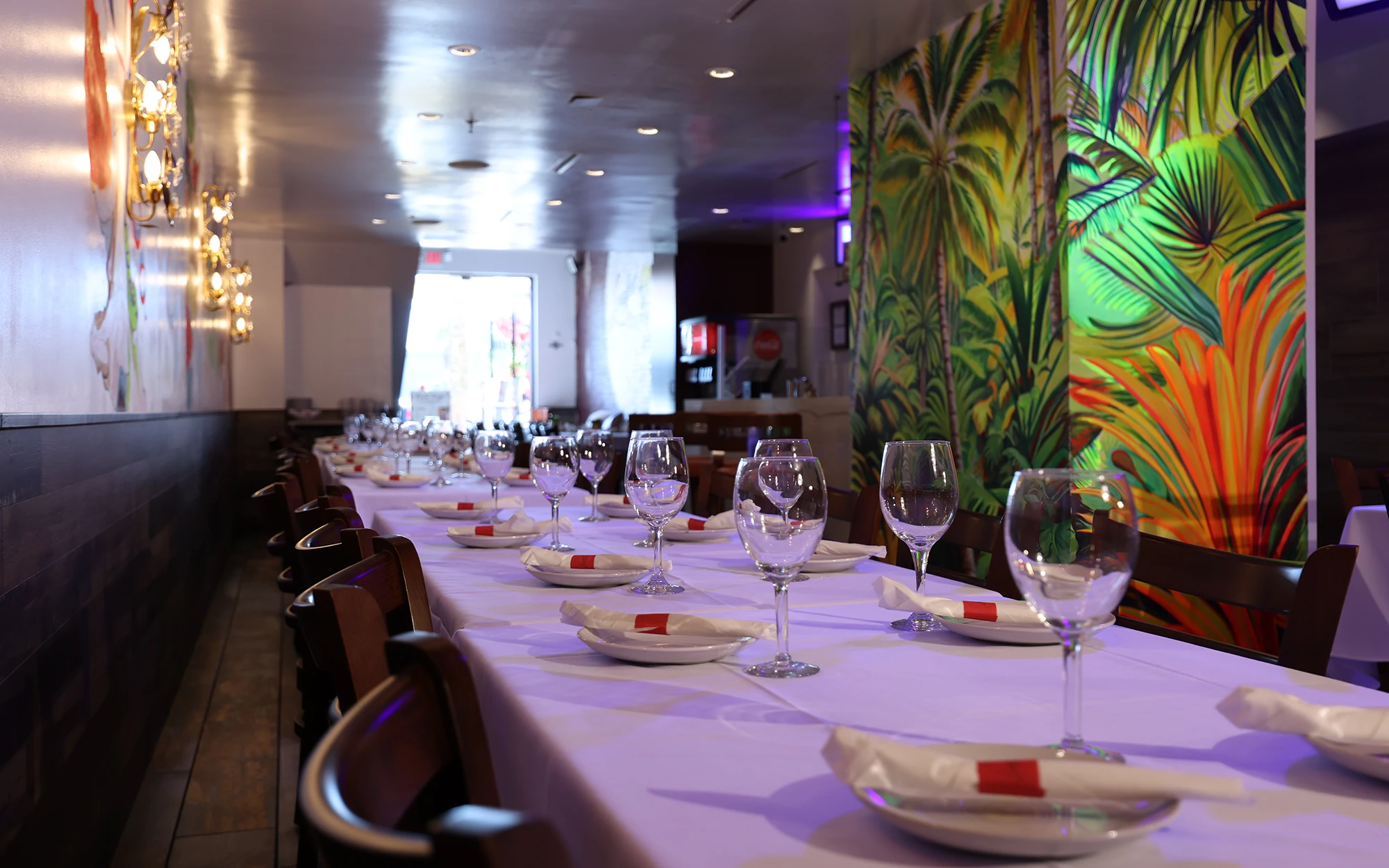 A long reserved section table set with white linens and wine glasses beside the tropical palm mural